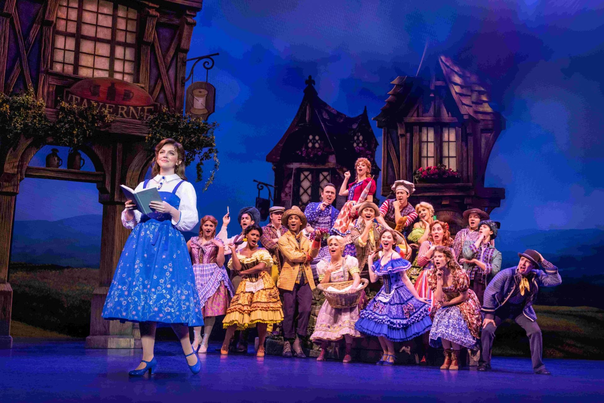 Stage musical cast in colourful village costumes sings behind woman reading a book.
