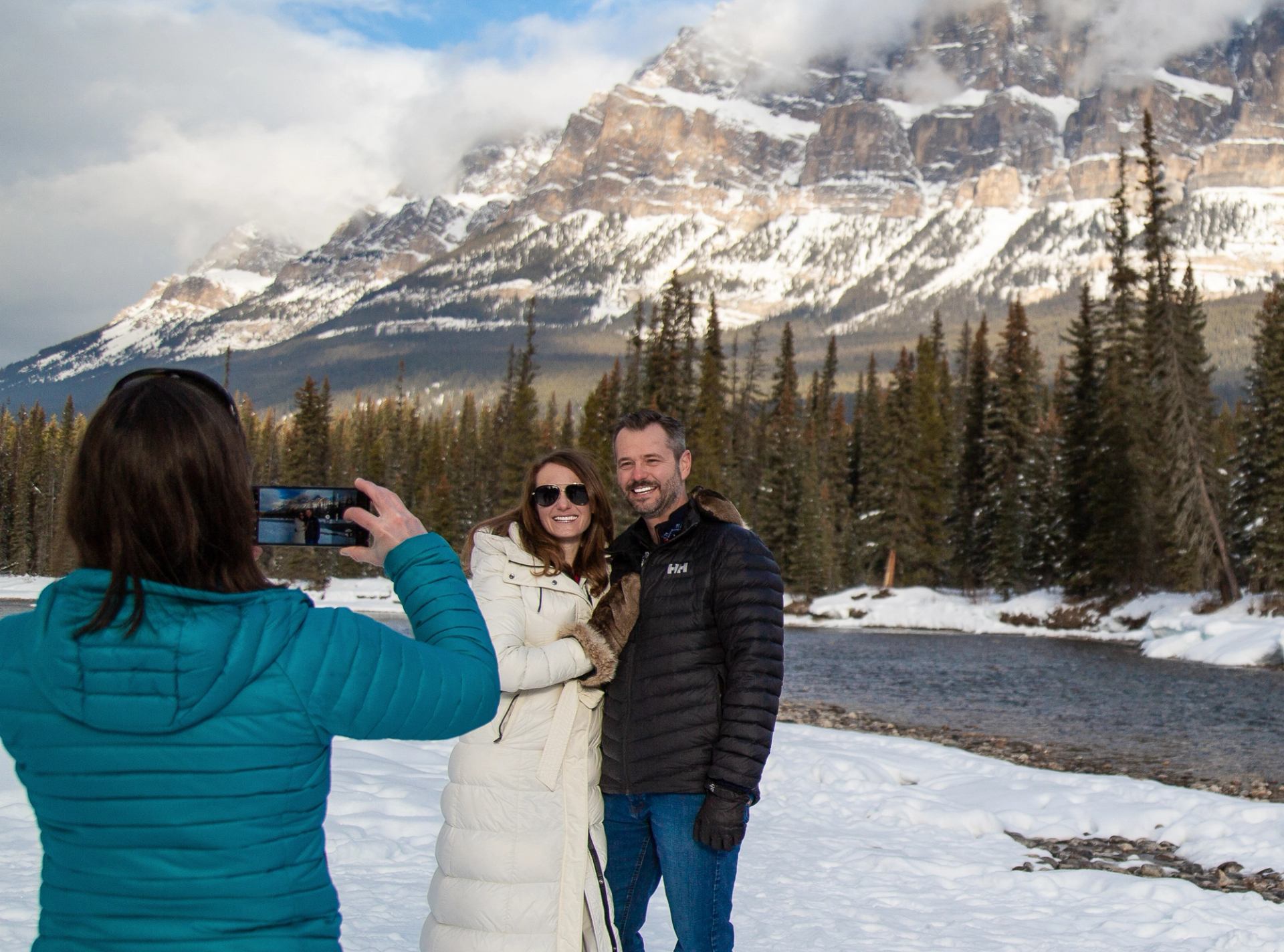 Person taking a photo of two people standing by a snowy river with mountains behind.