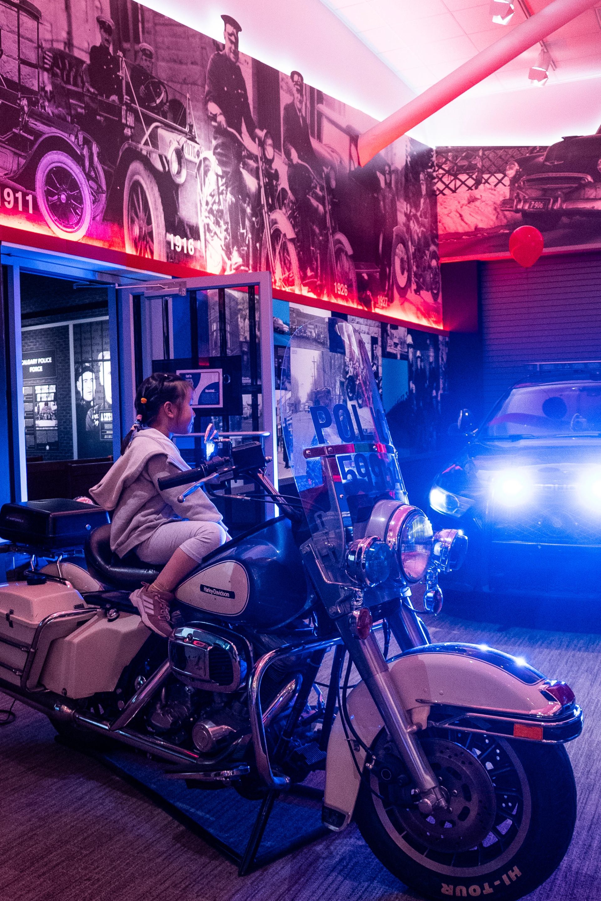 Child sits on police motorcycle in museum with vintage photos and patrol car.