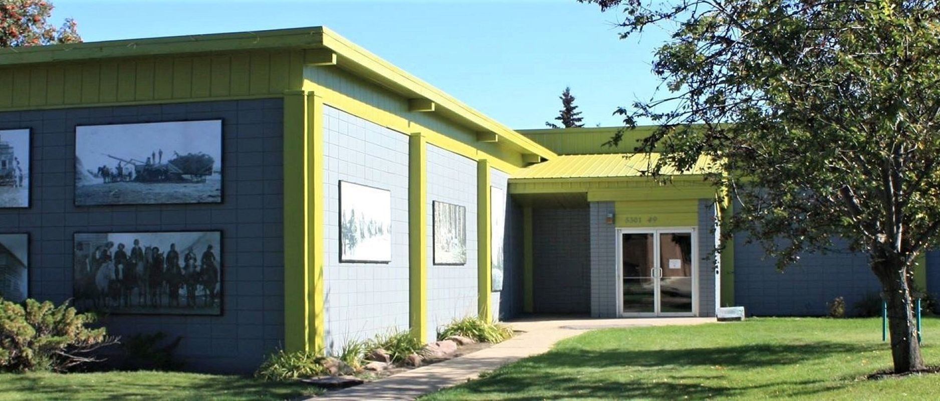 Single-storey grey building with yellow trim, outdoor photo murals, trees and grass.