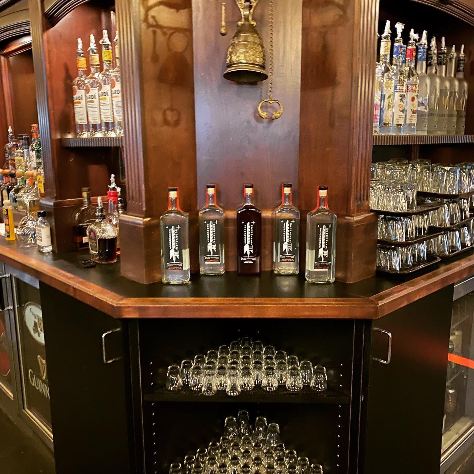 Bar counter with liquor bottles, stacked glassware, and a brass bell on wooden panel.