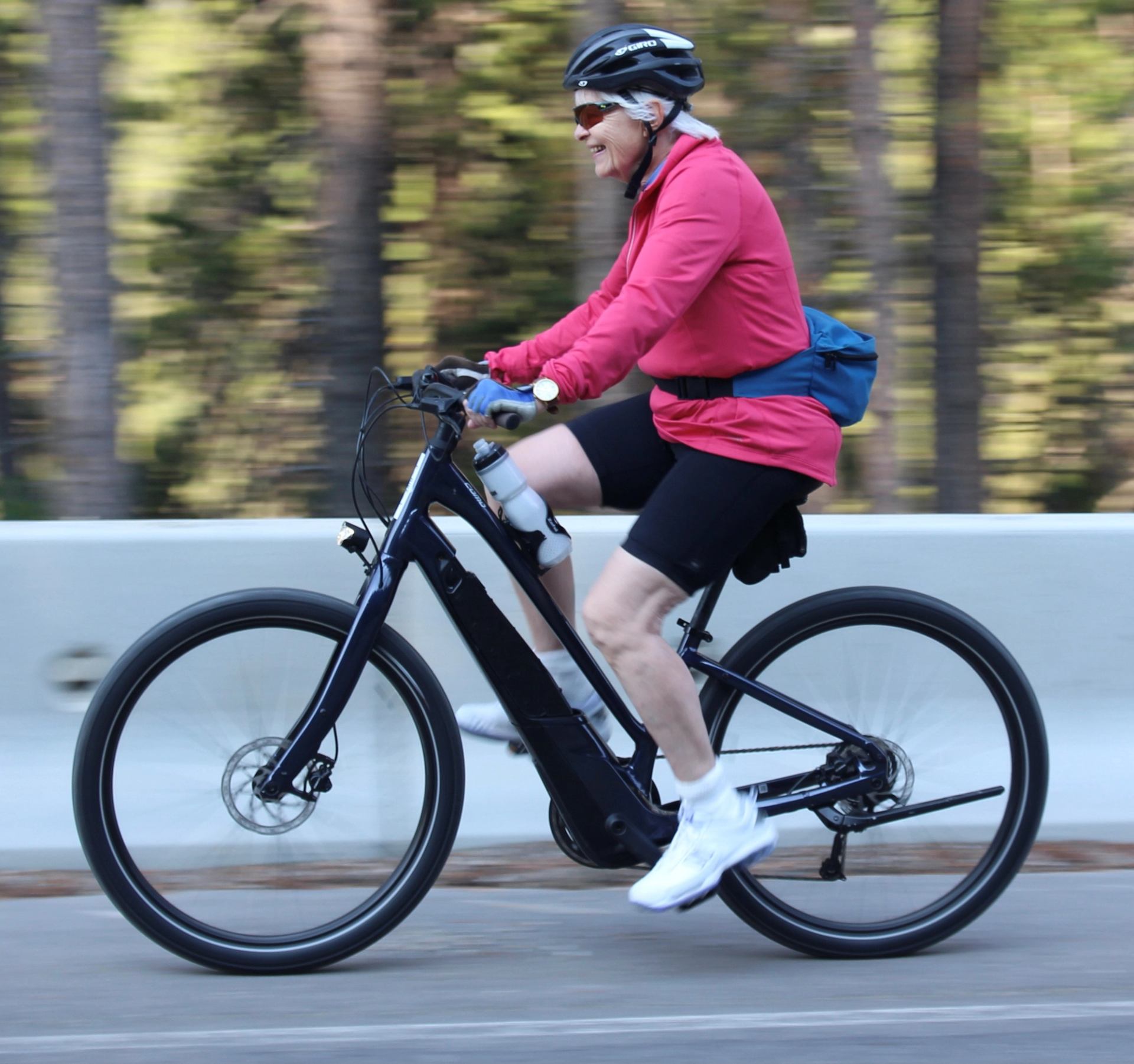 Cyclist in pink jacket riding on tree-lined road with concrete barrier.