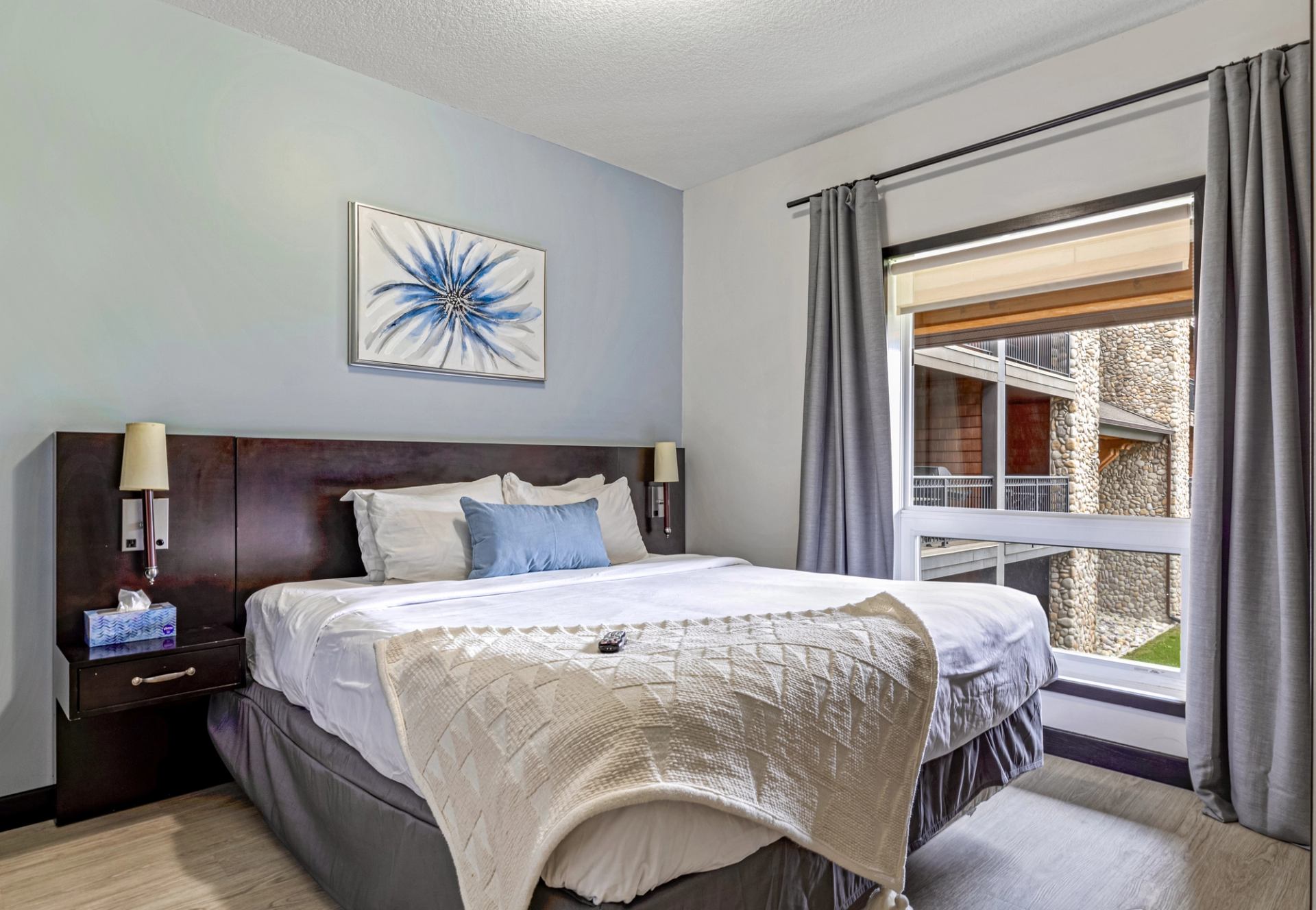 Bedroom with white bedding, blue accents, floral artwork and a large curtained window.