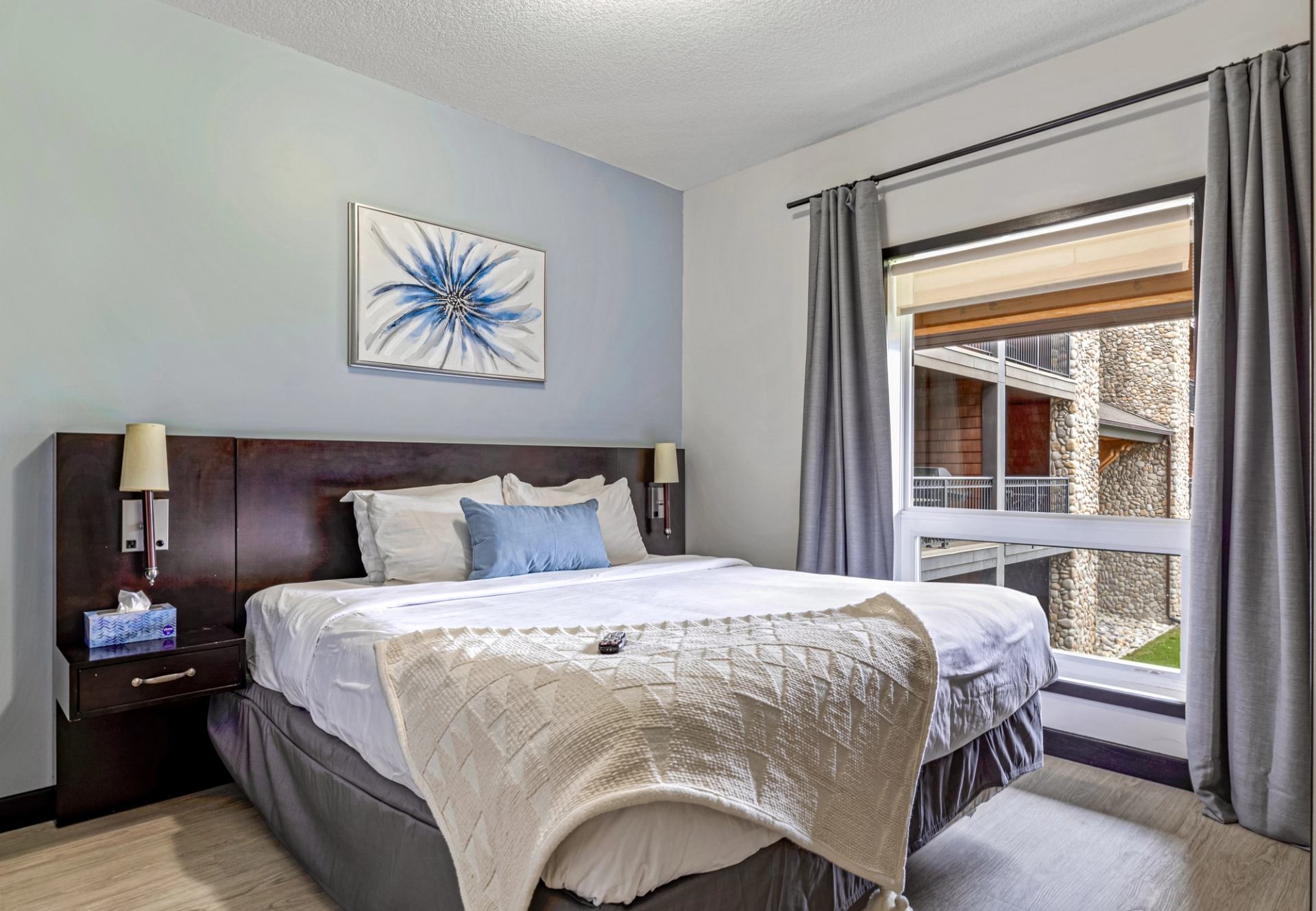 Bedroom with white bedding, blue accents, floral artwork and a large curtained window.