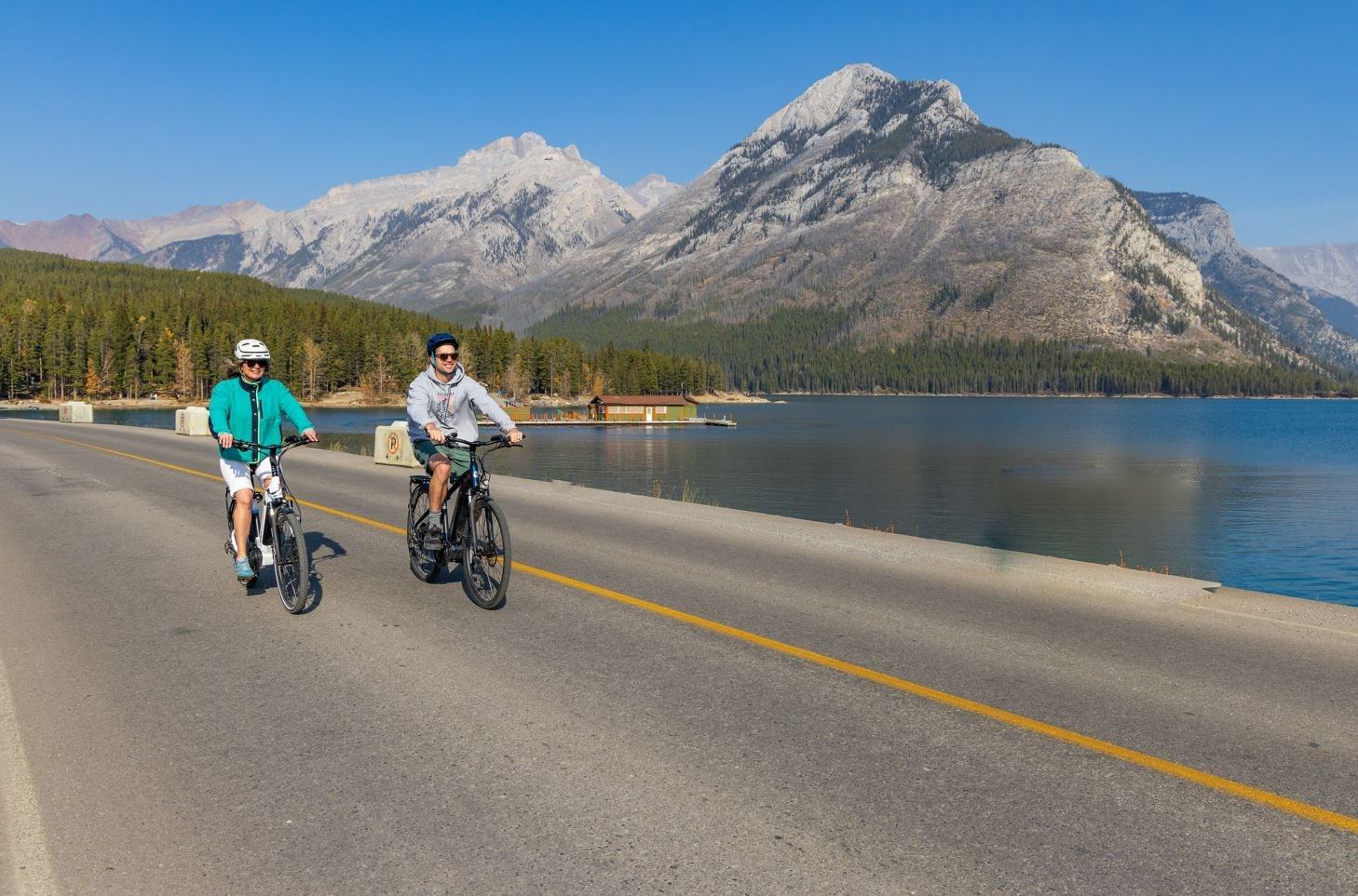 Two cyclists ride lakeside road with mountain views under clear skies in Banff.