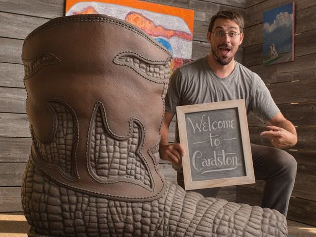 Person holding 'Welcome to Cowboytown' sign with cowboy boot and wooden backdrop.