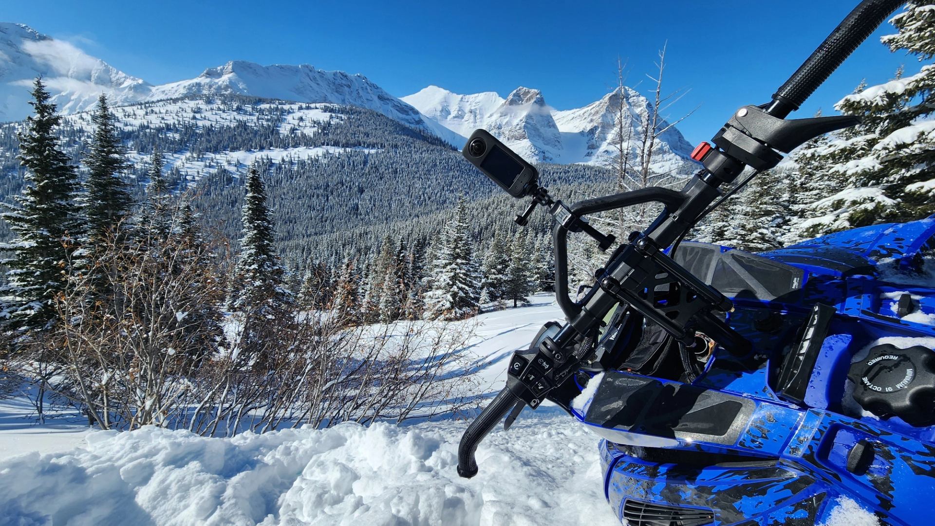 Blue snowmobile with handlebars and camera, overlooking snowy mountains and forest valley.