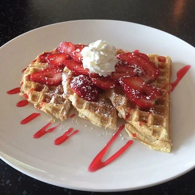 Waffle quarters with sliced strawberries, whipped cream and red sauce drizzle on white plate.