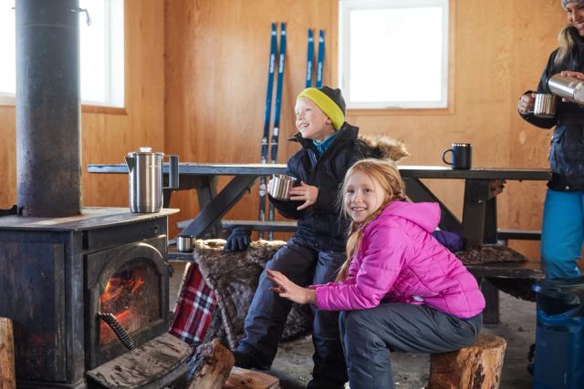 A family enjoys a hot drink while warming up around a fire, on a guided tour with Tamarack Outdoors in Waterton Lakes National Park