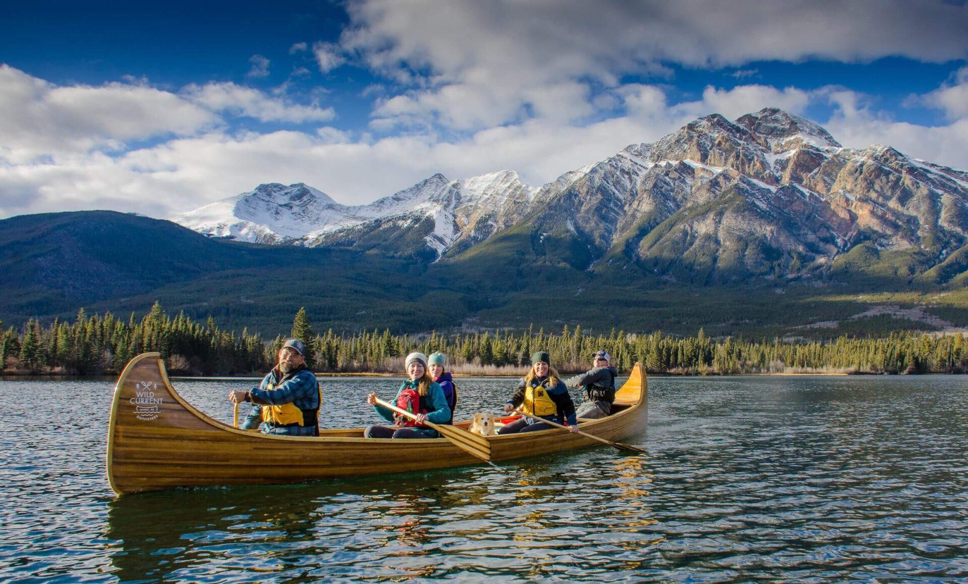 Group paddling a wooden canoe across a calm lake with evergreen forest and snow-capped mountains behind.