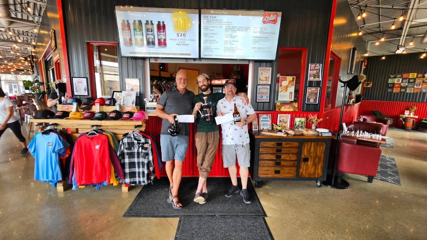 Three visitors pose inside a taproom surrounded by merchandise displays, seating areas, and menus.