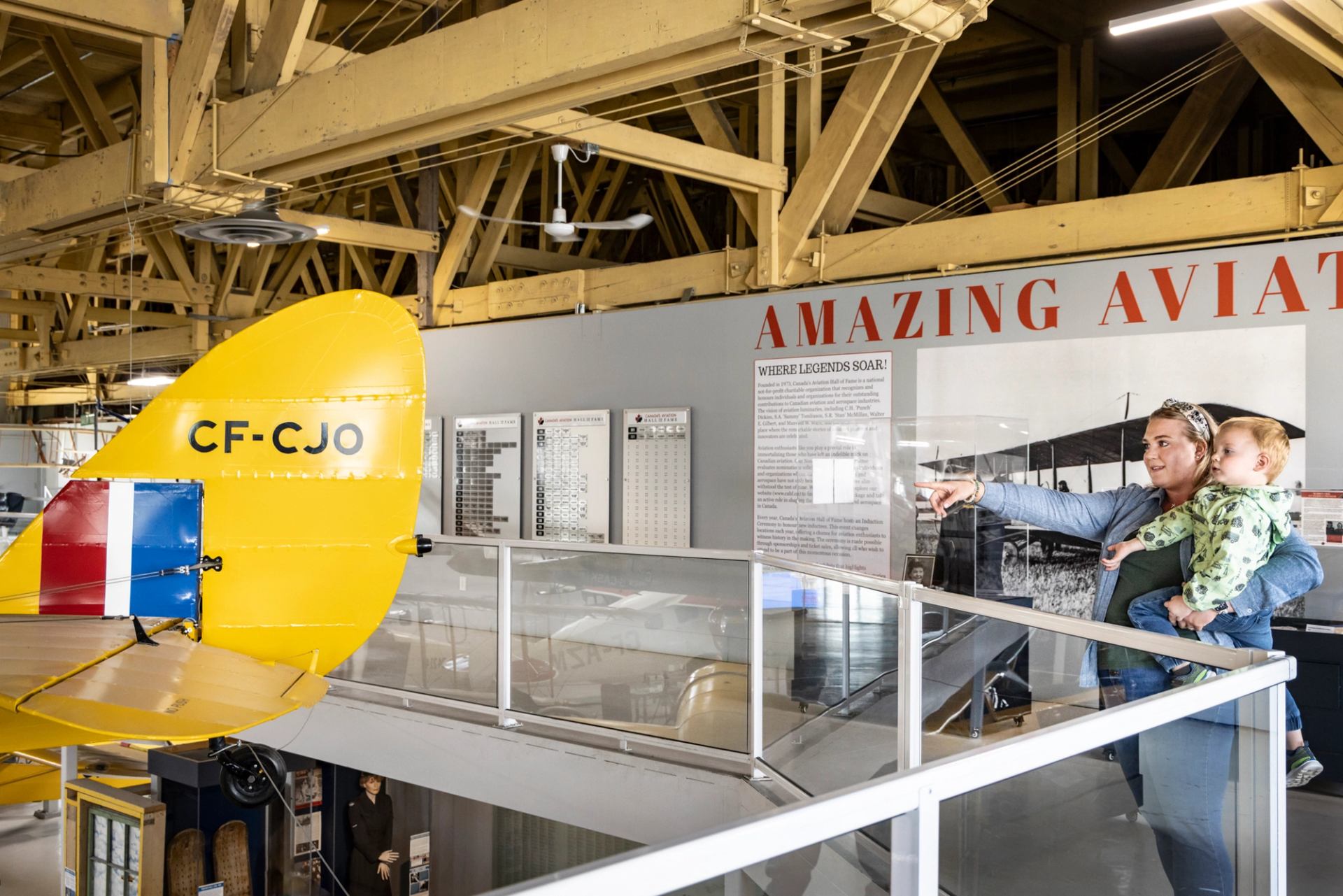 A woman holding a child points right in an aviation museum, with a yellow plane tail in the foreground and "AMAZING AVIATION" on the wall.