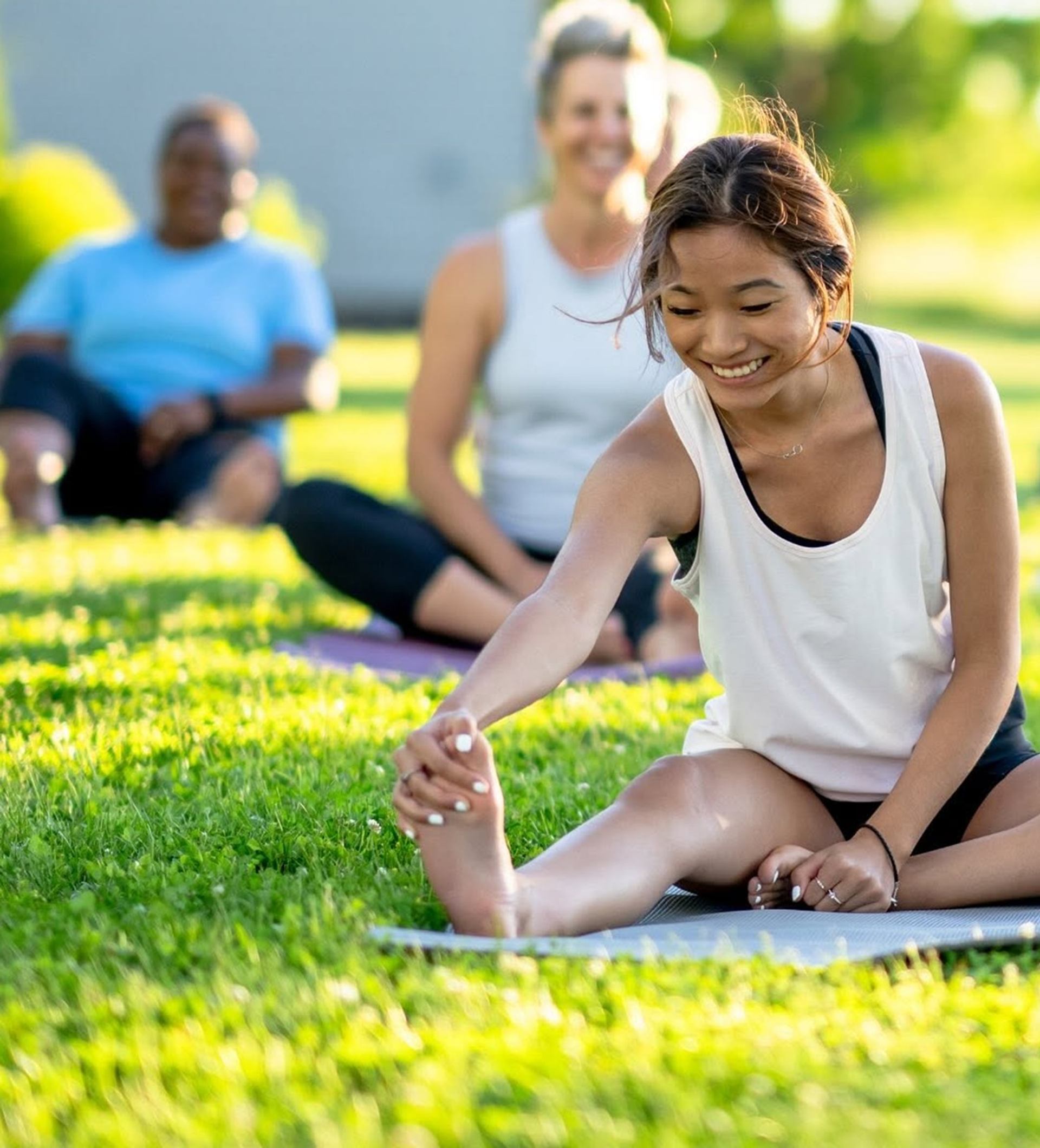 People on yoga mats in a park, person in front stretching one leg forward.