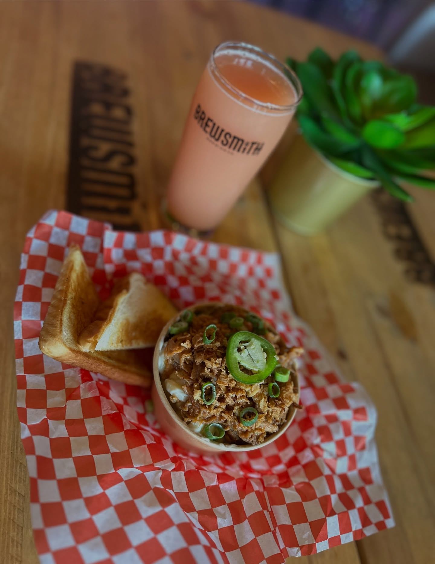 A pink craft beer sits beside toast and a bowl topped with crispy onions and jalapeño slices.