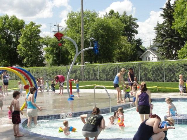 Children and adults enjoying shallow splash pool with water features on a sunny day.