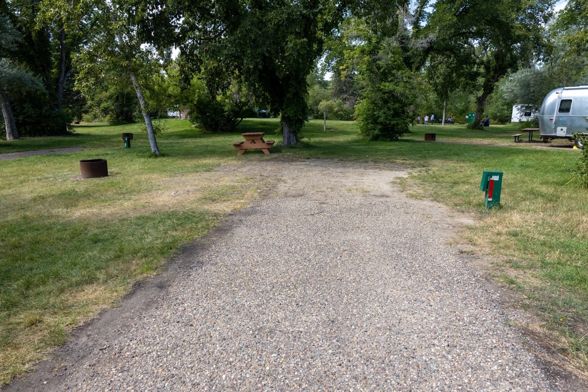 A gravel campsite with a picnic table, fire pit, utility hook-up, and grassy areas under trees.