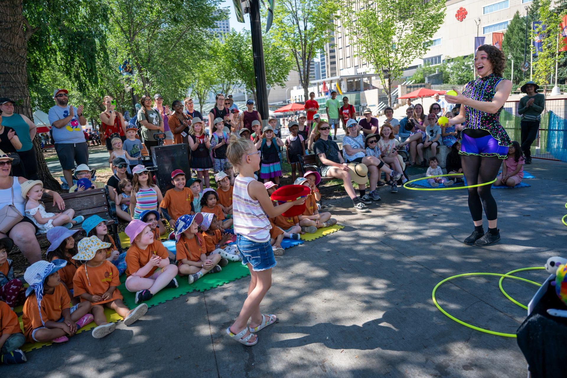 Street performer juggles for seated children and adults watching in a shaded urban park.