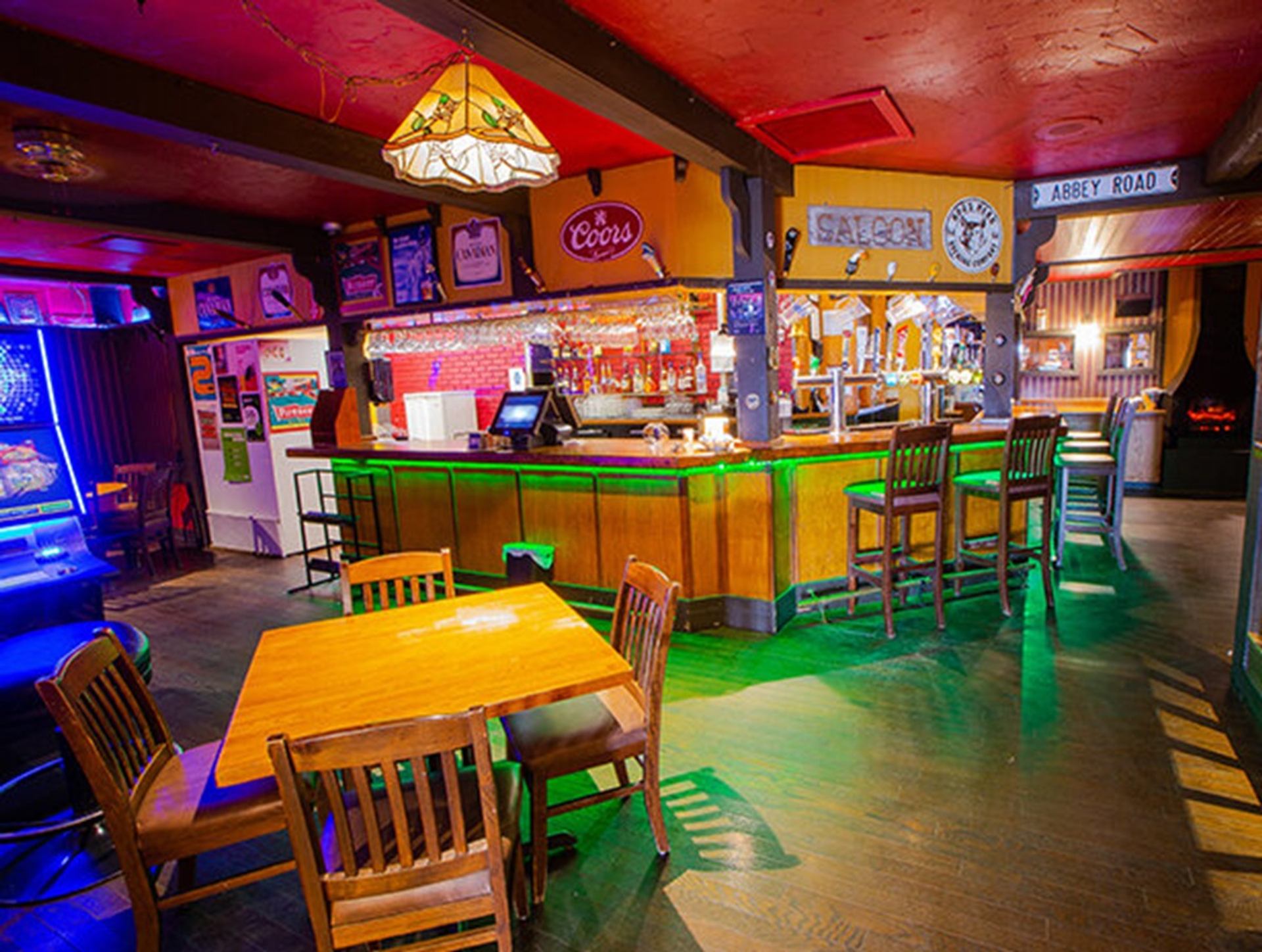 A cozy bar area features a wooden counter with stools, colorful accent lighting, and signage on the walls. Tables and chairs sit in the foreground, while gaming machines and warm lighting create a casual pub-style atmosphere.