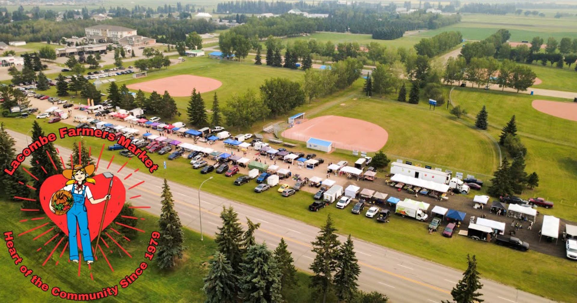 An aerial view shows market vendors, vehicles, sports fields, trees, and surrounding green spaces.