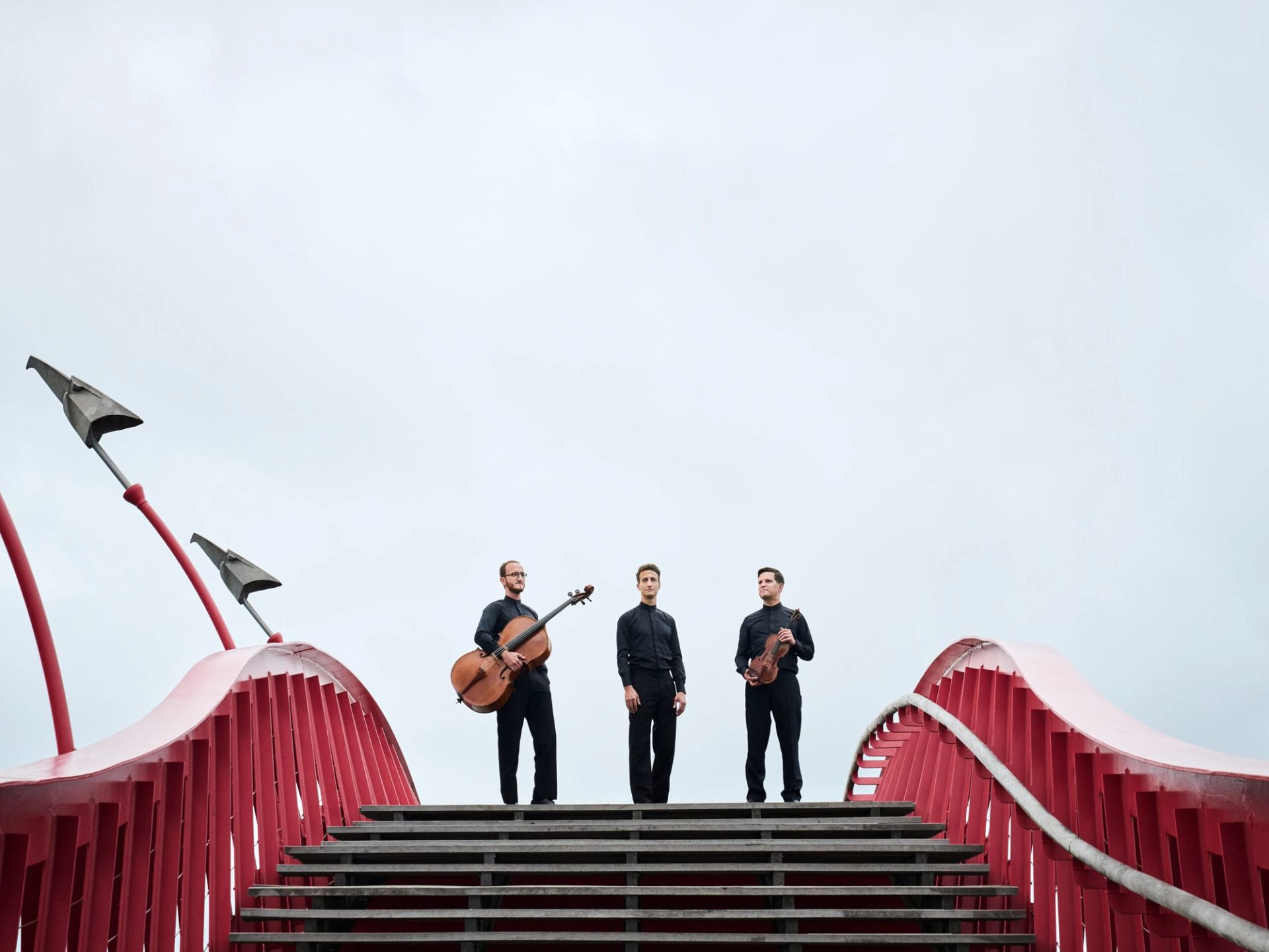 Three musicians with string instruments stand on red arched bridge against cloudy sky.