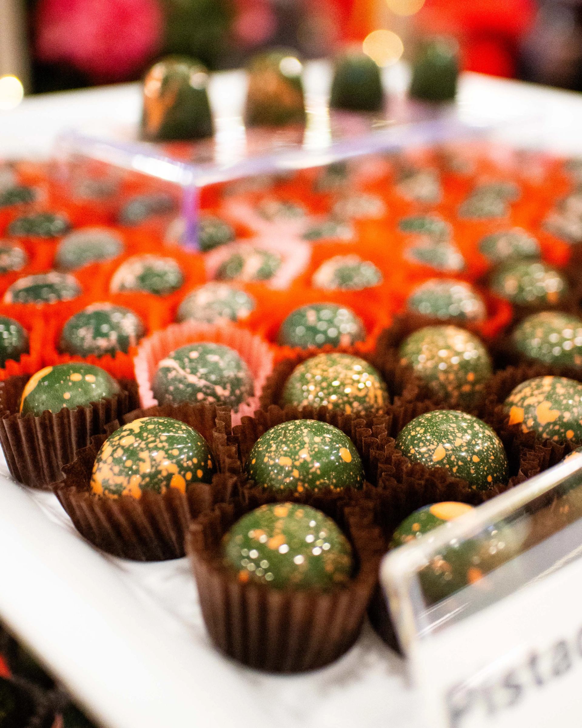 Rows of green speckled chocolate truffles in brown and orange paper cups on display.
