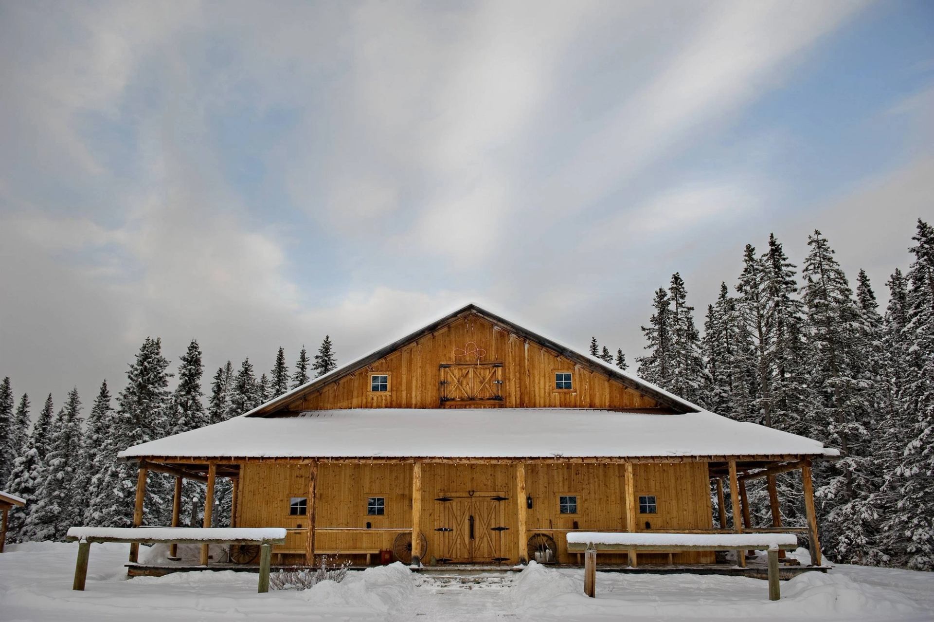 Snowy barn in countryside surrounded by trees under a partly cloudy sky.