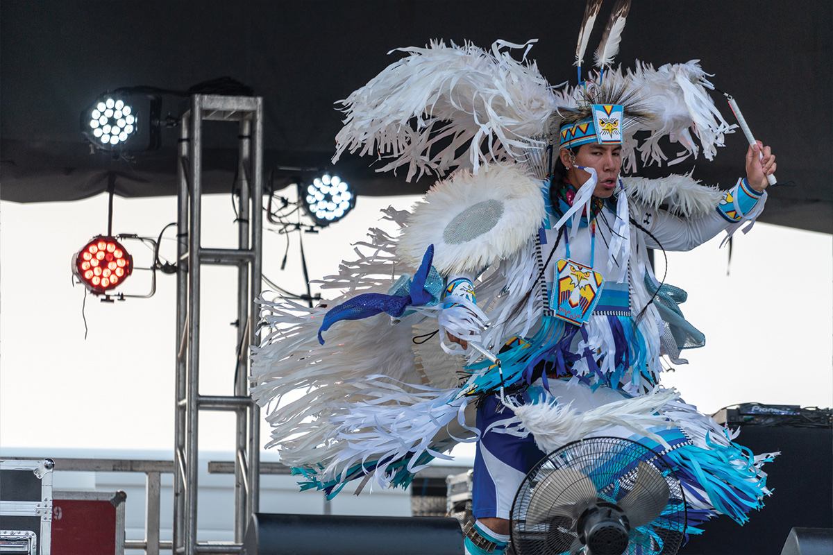 Powwow dancer in blue and white regalia performs on stage under concert lights.