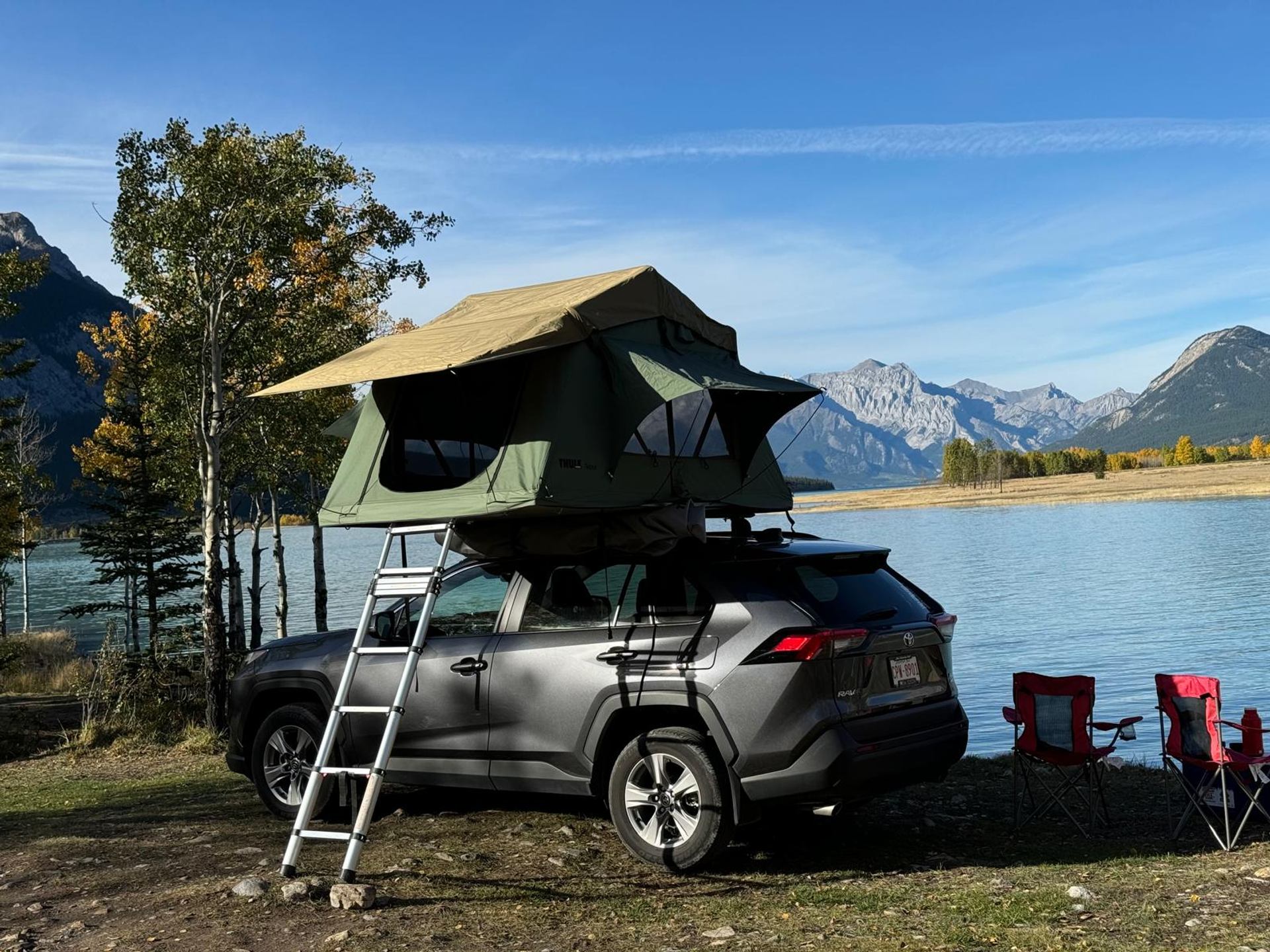 SUV with rooftop tent parked by lake, mountains and trees in background, camping chairs nearby.