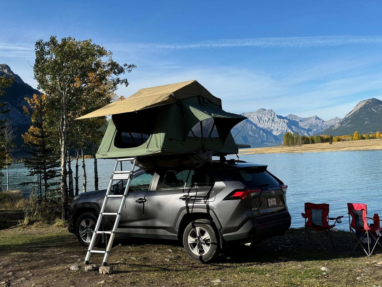 SUV with rooftop tent parked by lake, mountains and trees in background, camping chairs nearby.