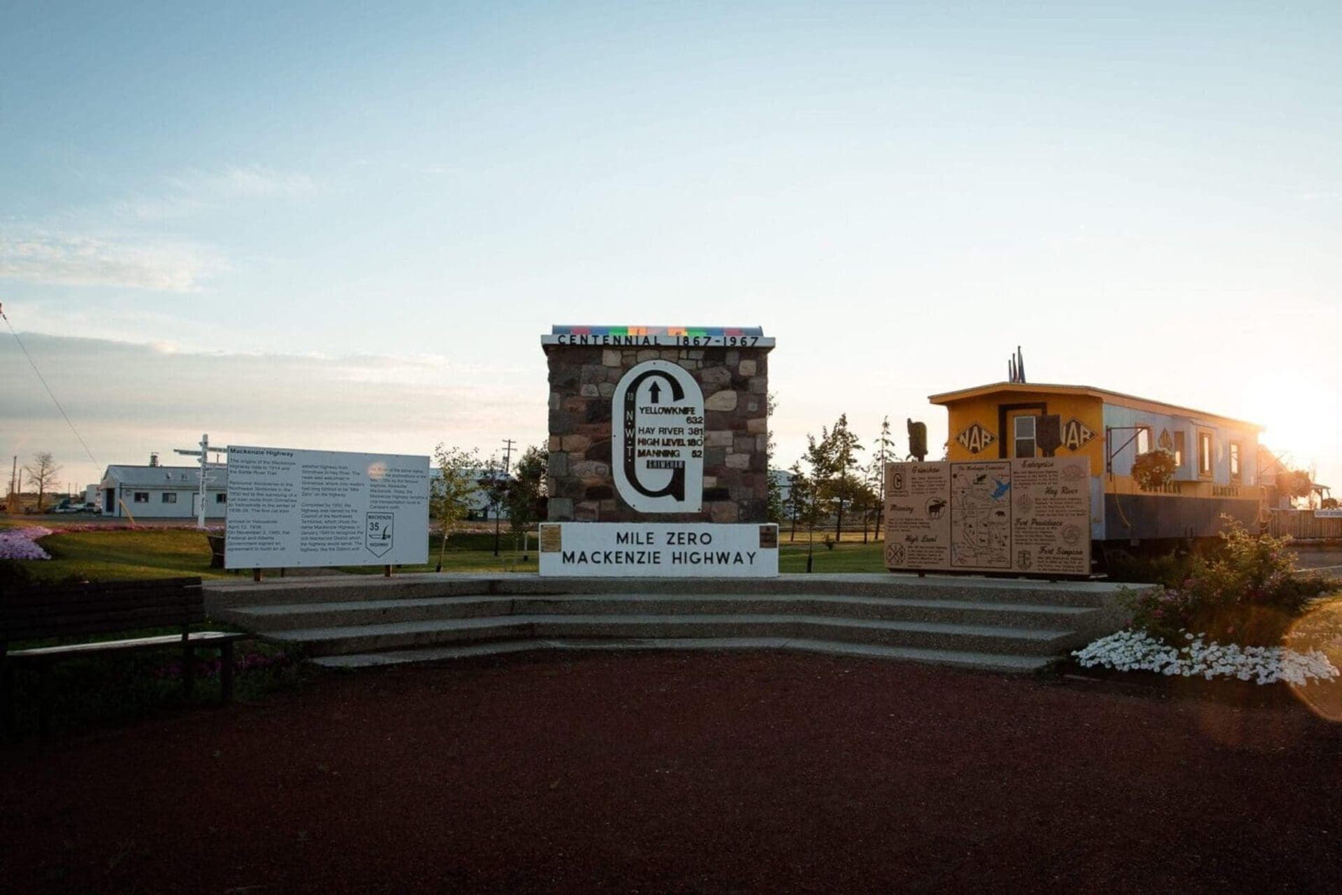 Mackenzie Highway Mile Zero monument with stone pillar, signs and yellow railcar at sunset.