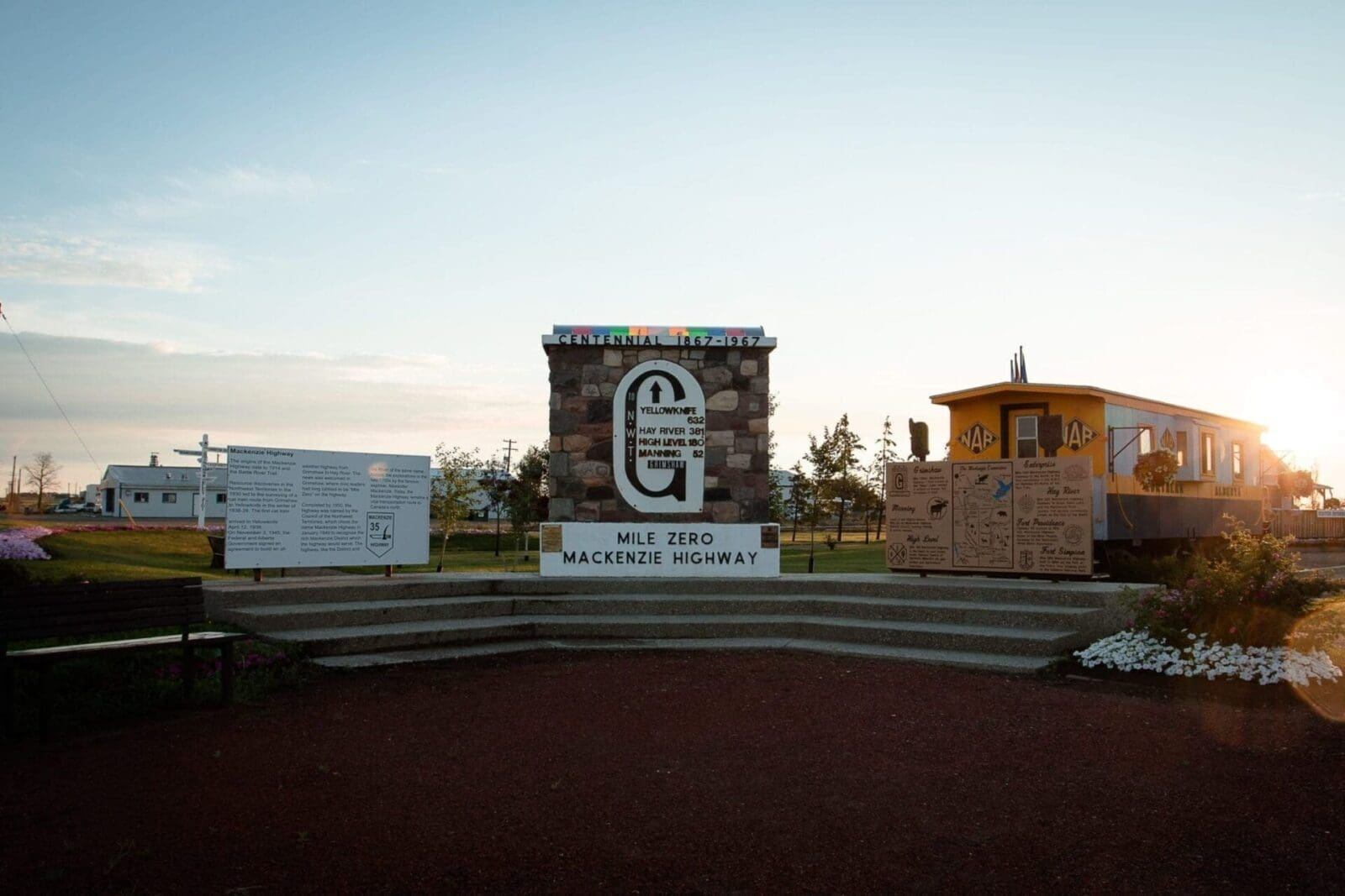 Mackenzie Highway Mile Zero monument with stone pillar, signs and yellow railcar at sunset.