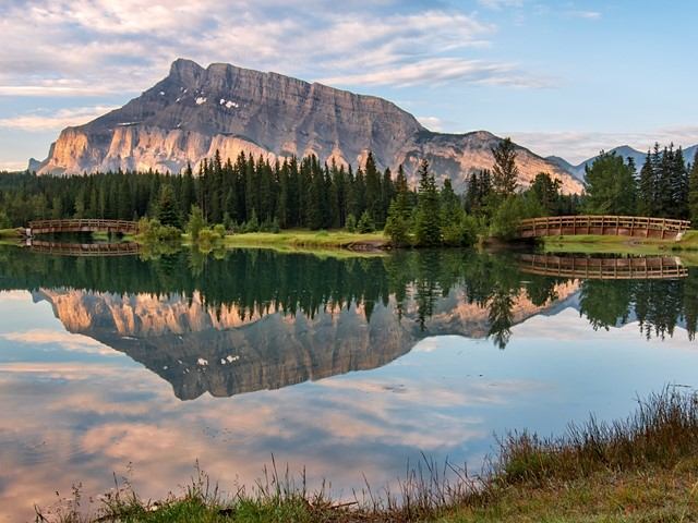 Lakeside reflection of mountains with evergreen trees and a wooden fence along the shore.
