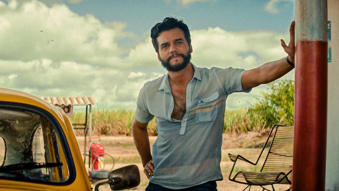 Man leaning against a post beside a yellow car at a rural roadside stop under a bright, open sky.