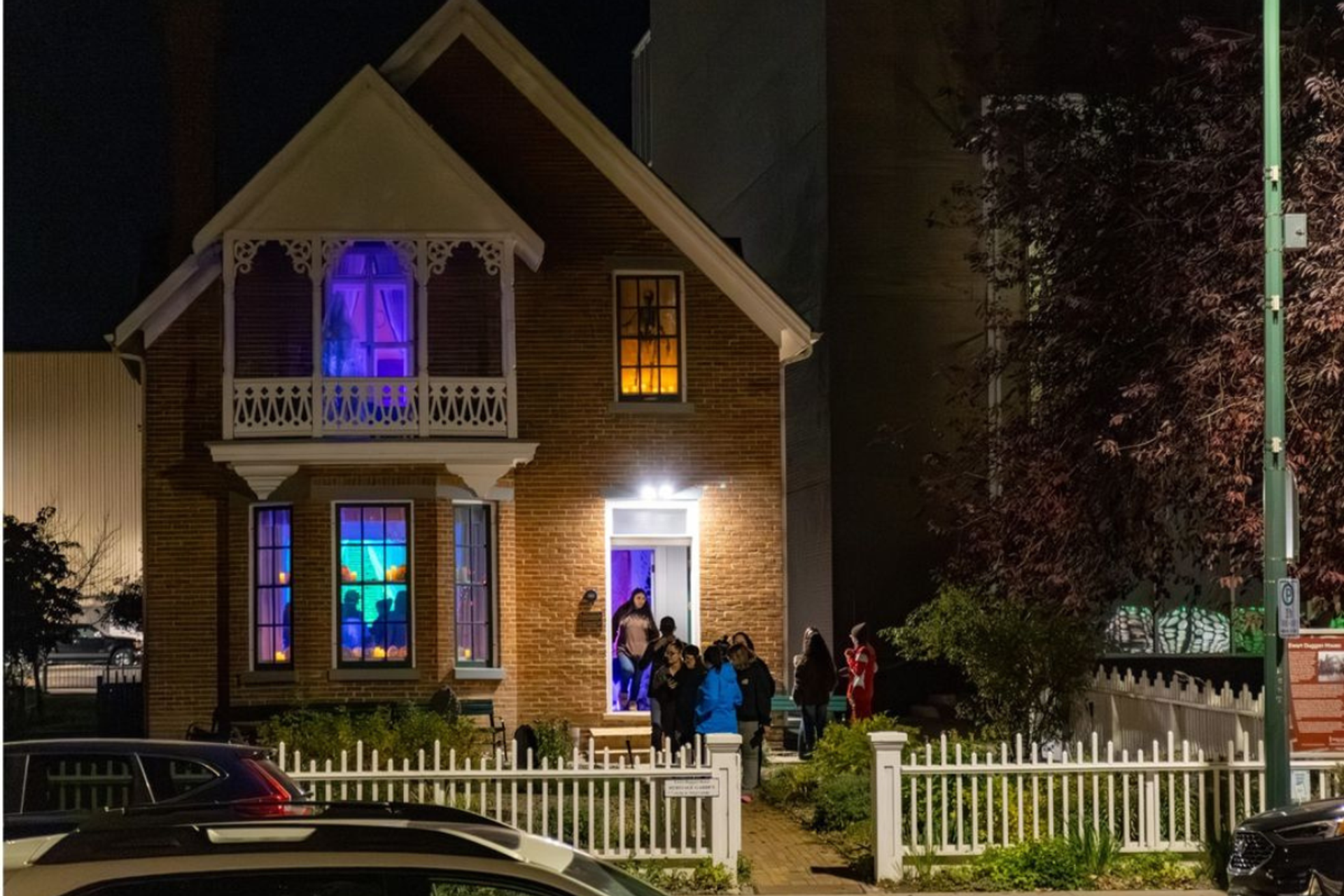 People gather outside a brick house with colourful illuminated windows at night.