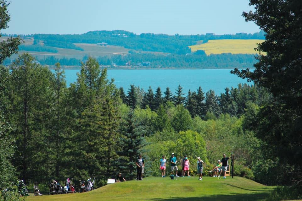 People gathered on grassy area with trees and water at Gull Lake Golf Course.