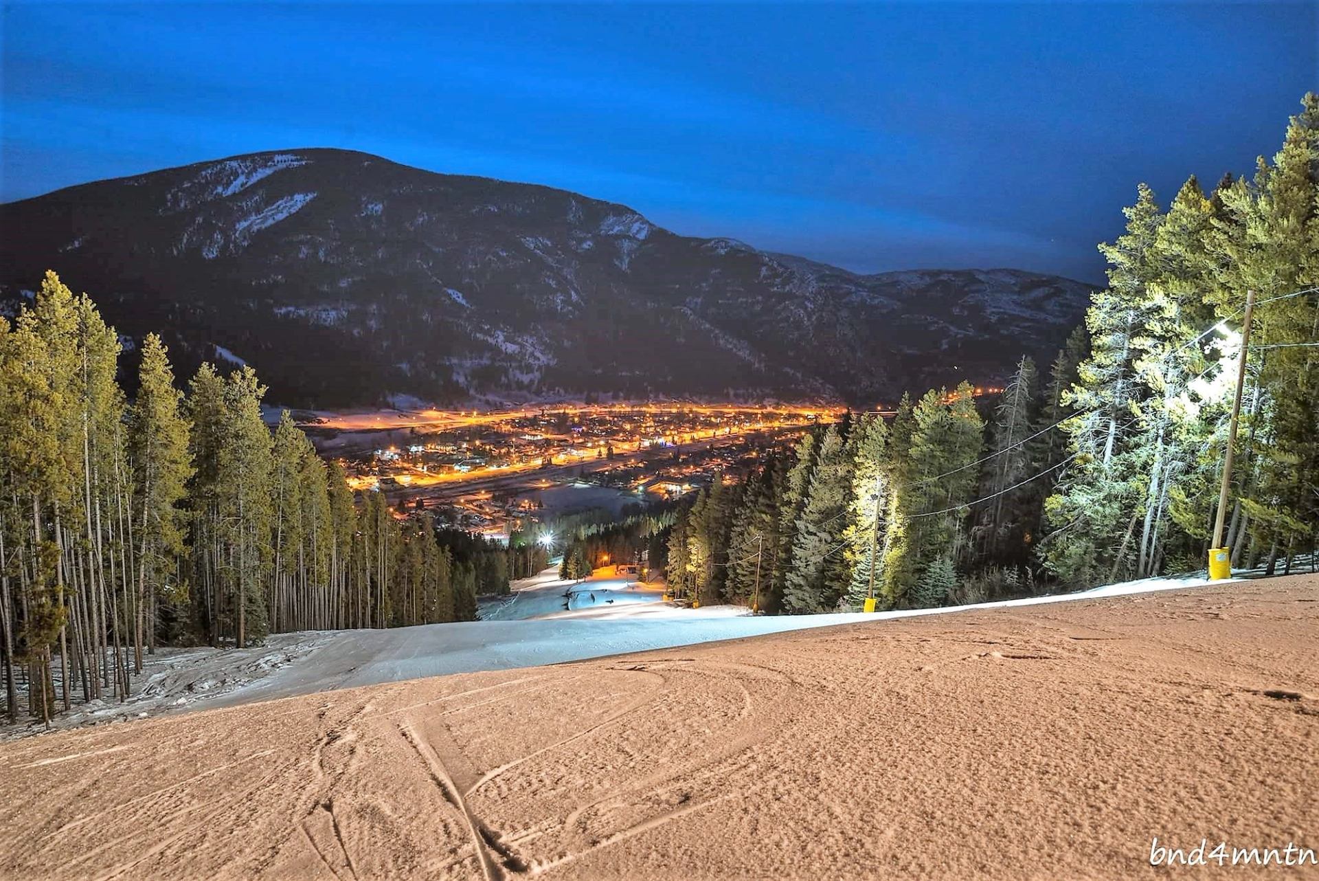 Lit ski run at night overlooking a glowing valley, framed by snow-covered trees and dark mountain slopes.