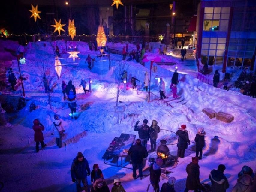 People gather around lights and fire pits in snowy night at winter celebration.