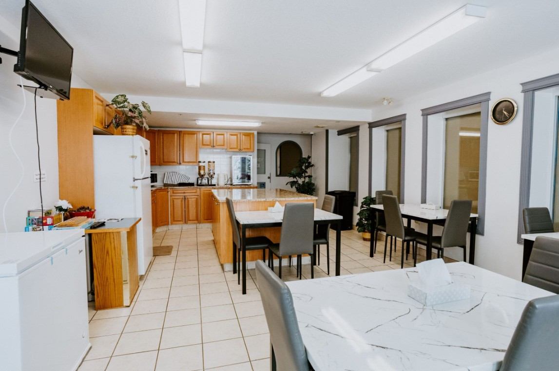 Bright communal kitchen with wooden cabinets, marble tables, grey chairs and wall-mounted television.