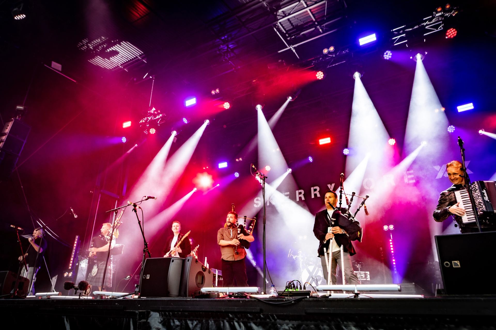 Band performs on stage with bagpipes and accordion under colourful concert lights.