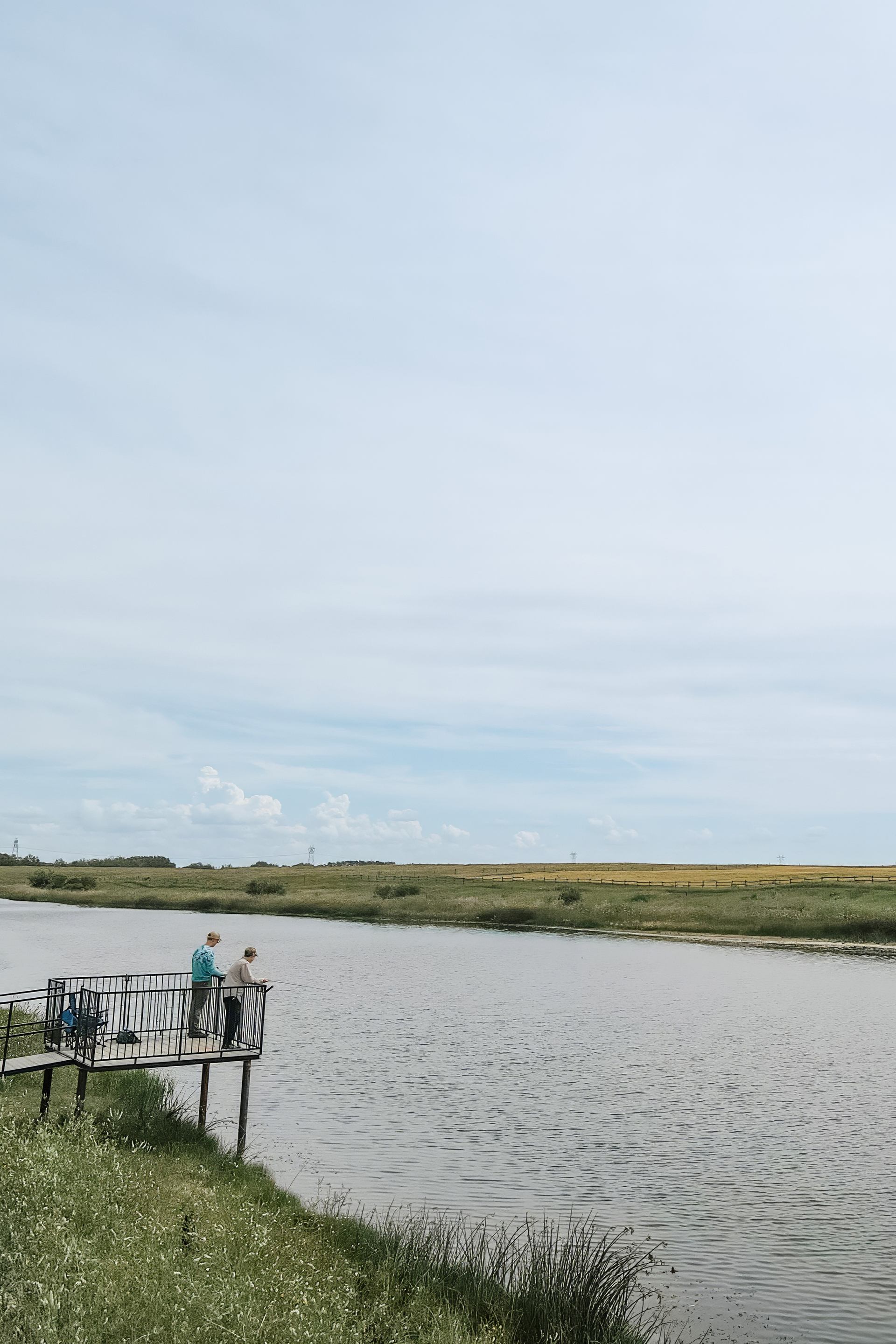 Two people stand on a small metal dock beside a calm rural river.