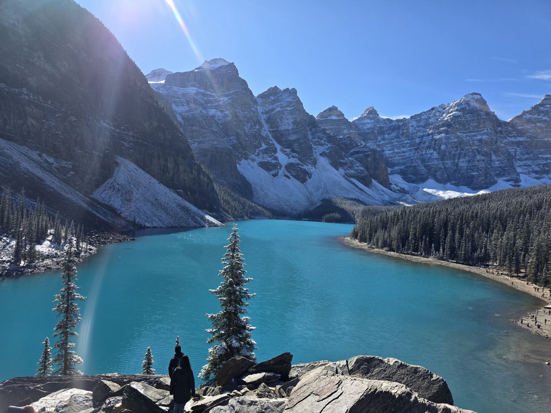 A vibrant turquoise lake nestled among snow-capped mountains and pine forests under a sunny sky, with a person in the foreground.