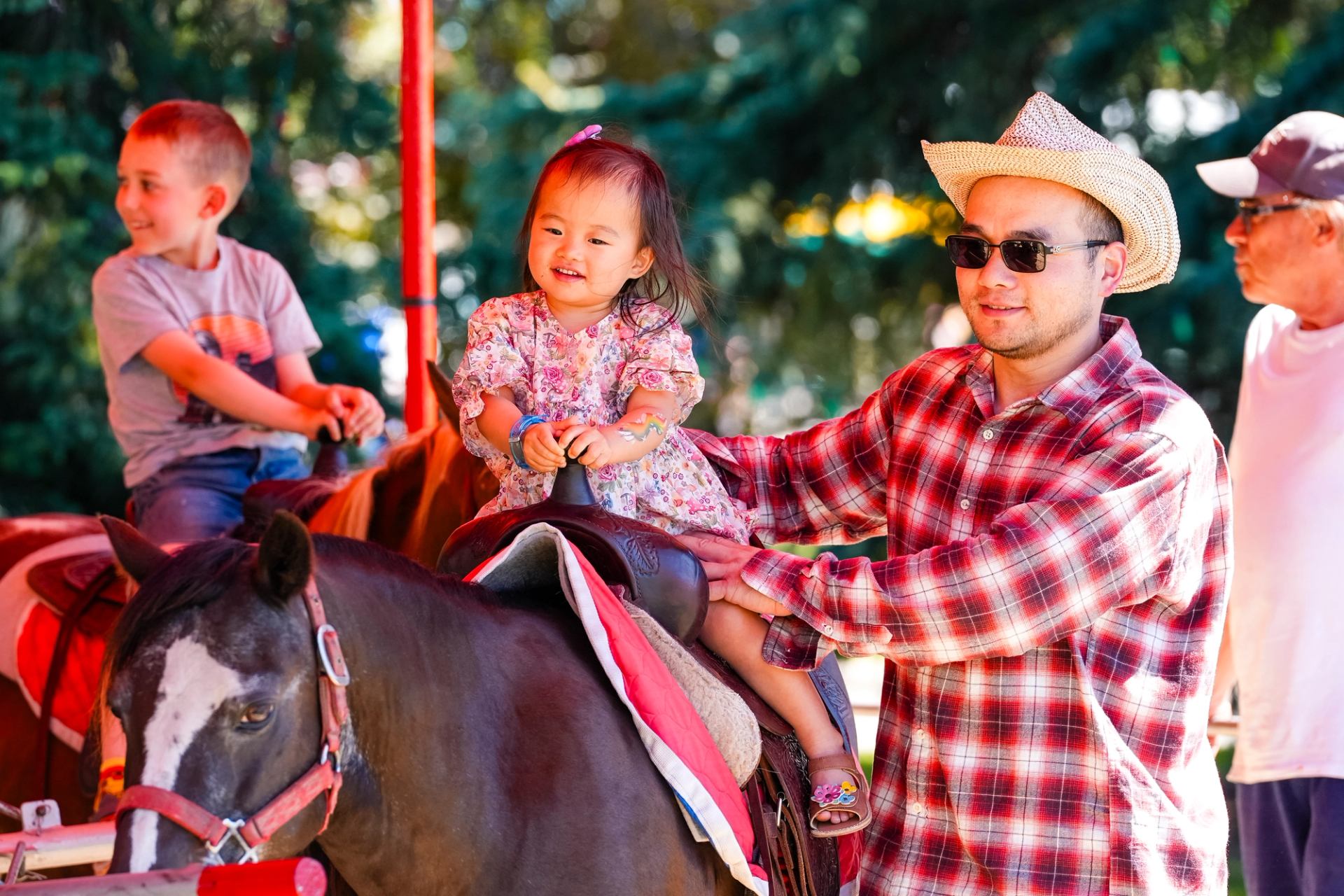 Children ride ponies with adults assisting at a shaded pony ride area.