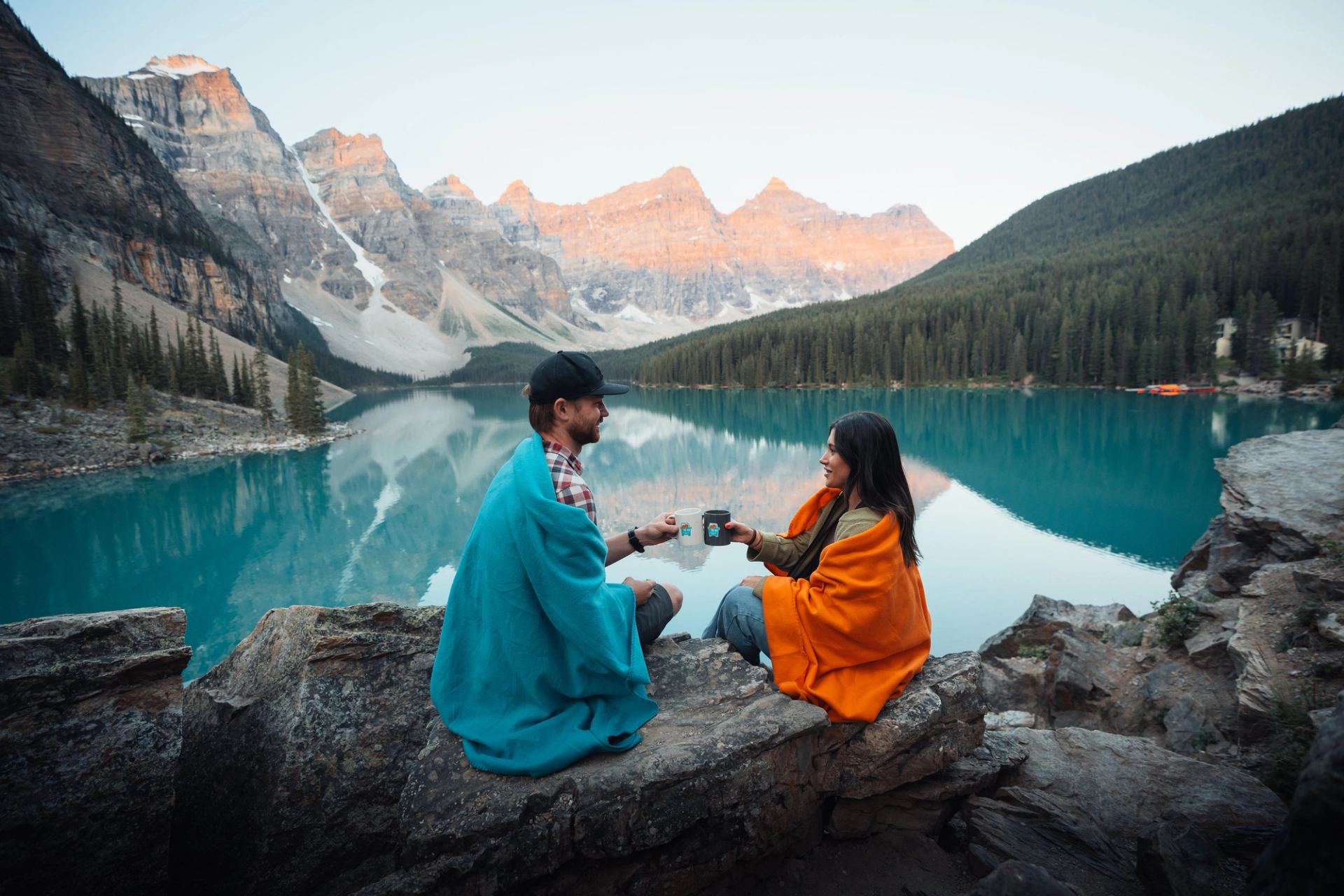 Two people wrapped in blankets sit by Moraine Lake with mountains and turquoise water.