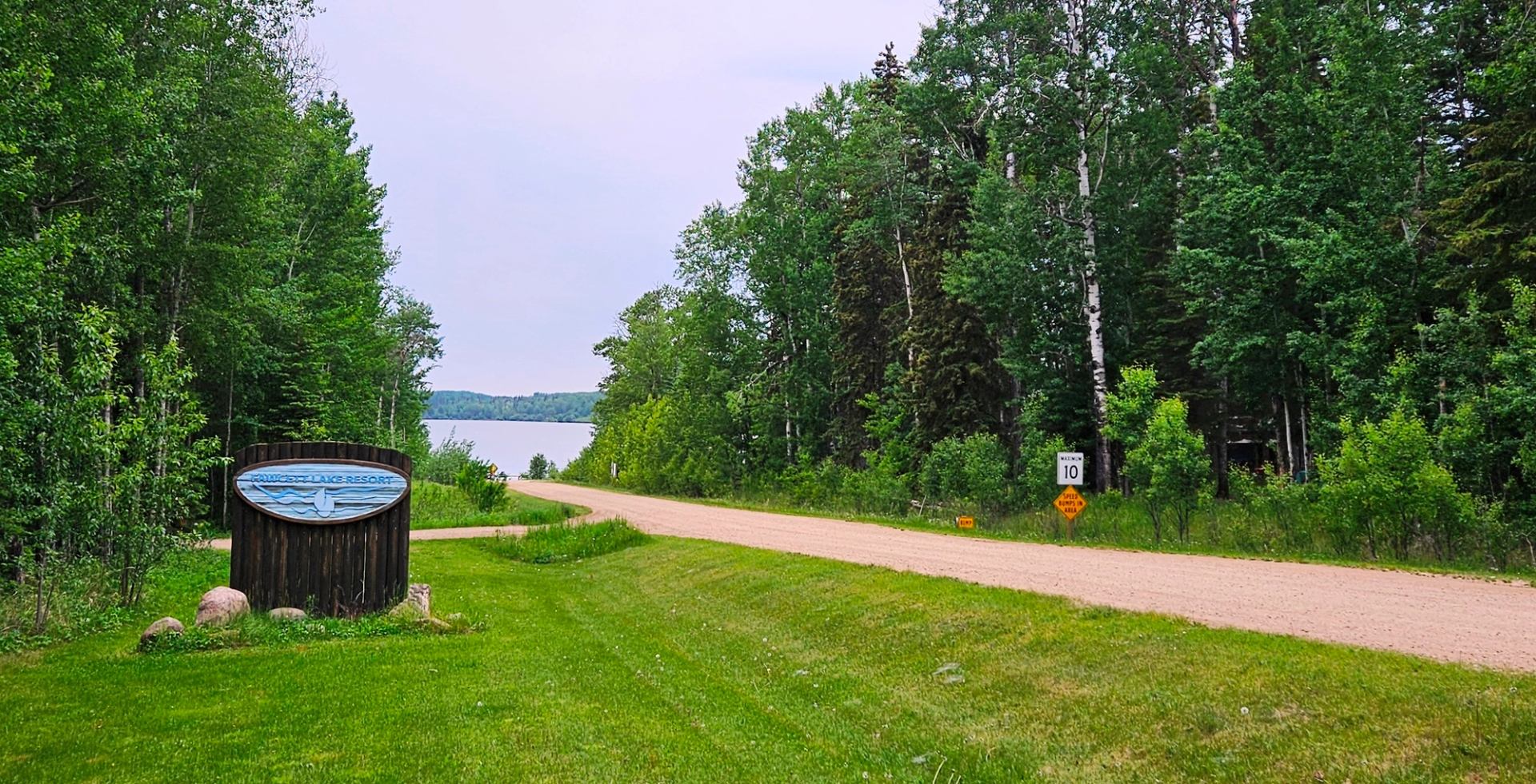 Gravel road leading to a lake, with resort sign and speed limit signs beside trees