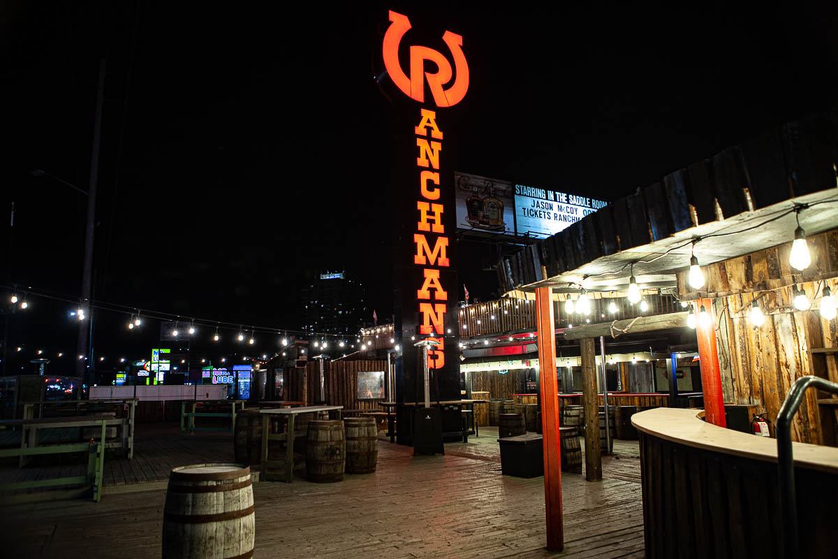 Outdoor Ranchman’s bar patio at night with string lights, barrels and wooden tables.