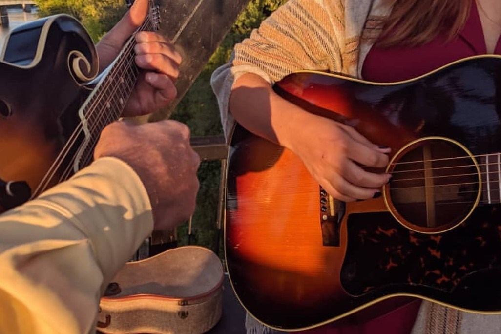 Guitarists playing acoustic and electric guitars with fingers on strings outdoors in daylight.