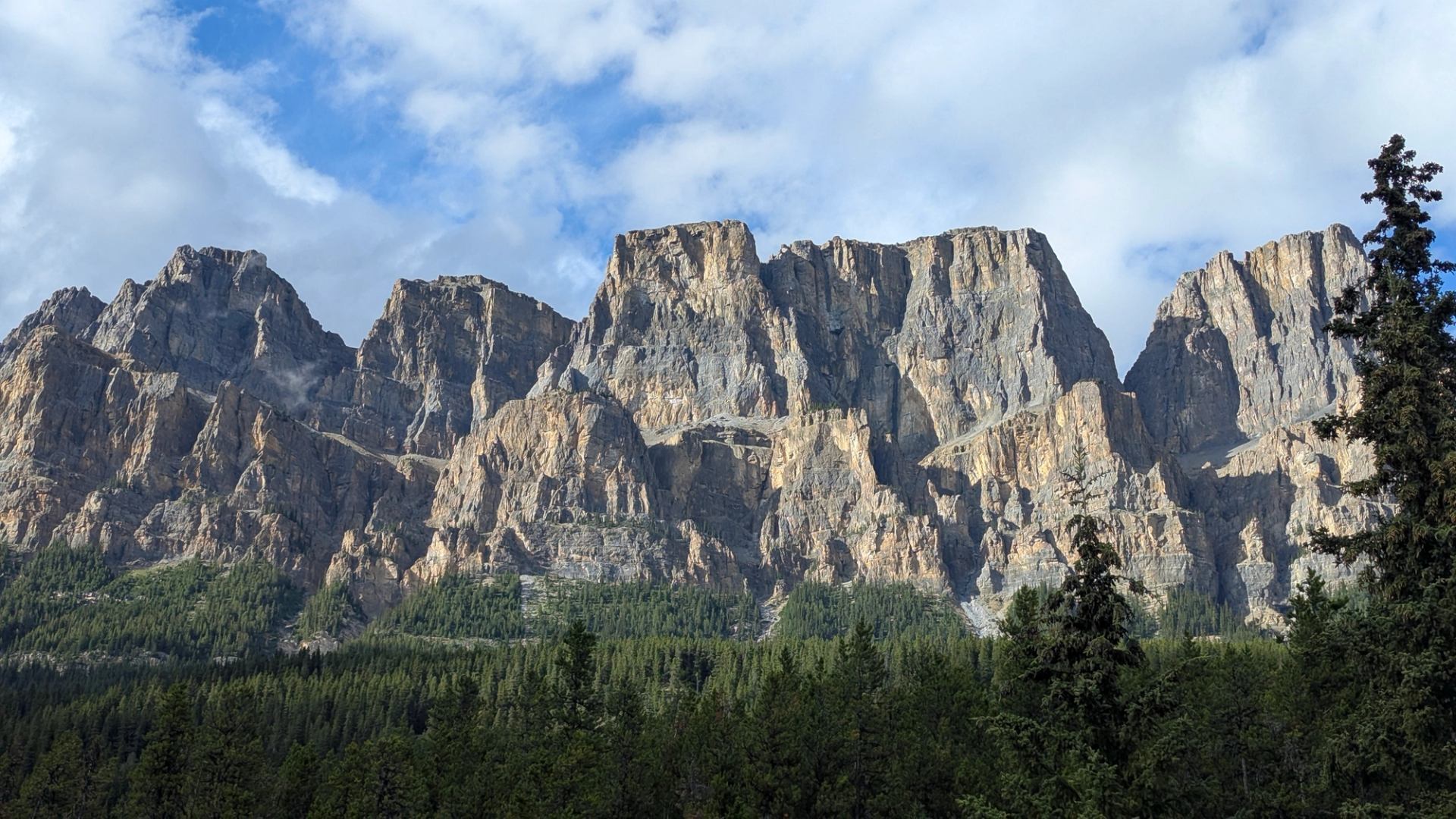 Rocky mountain cliffs above dense forest under a partly cloudy sky.
