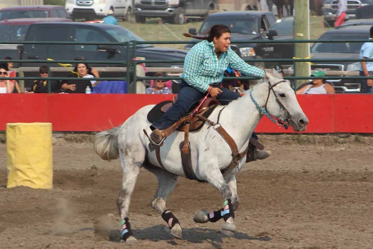 Rodeo rider on galloping white horse passes yellow barrel before spectators in stands.