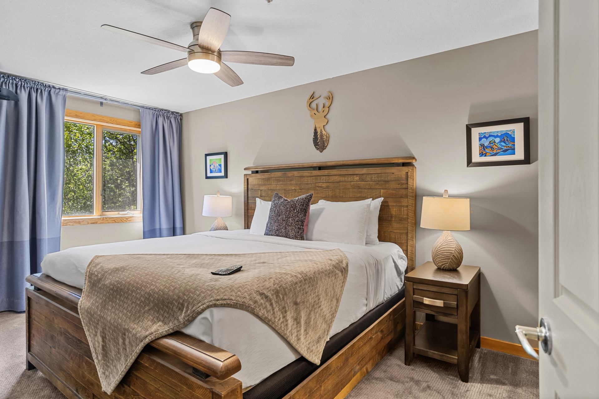 Bedroom with wooden bed, white bedding, two lamps, ceiling fan, and window with forest view.