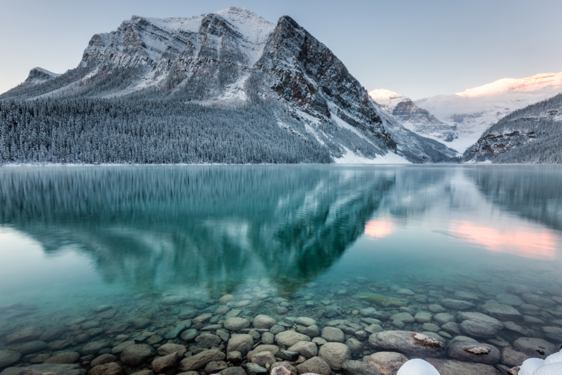 Snowy mountains reflected in a turquoise lake with clear rocks visible under the water.